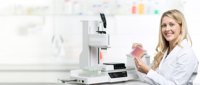 Scientist holds a 96 well plate, with the ASSIST PLUS pipetting robot in the background.