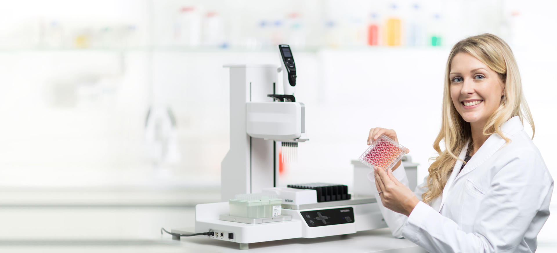 Scientist holds a 96 well plate, with the ASSIST PLUS pipetting robot in the background.