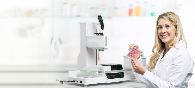 96ウェルプレートを手に持つ科学者と、その後ろのASSIST PLUSピペティングロボット